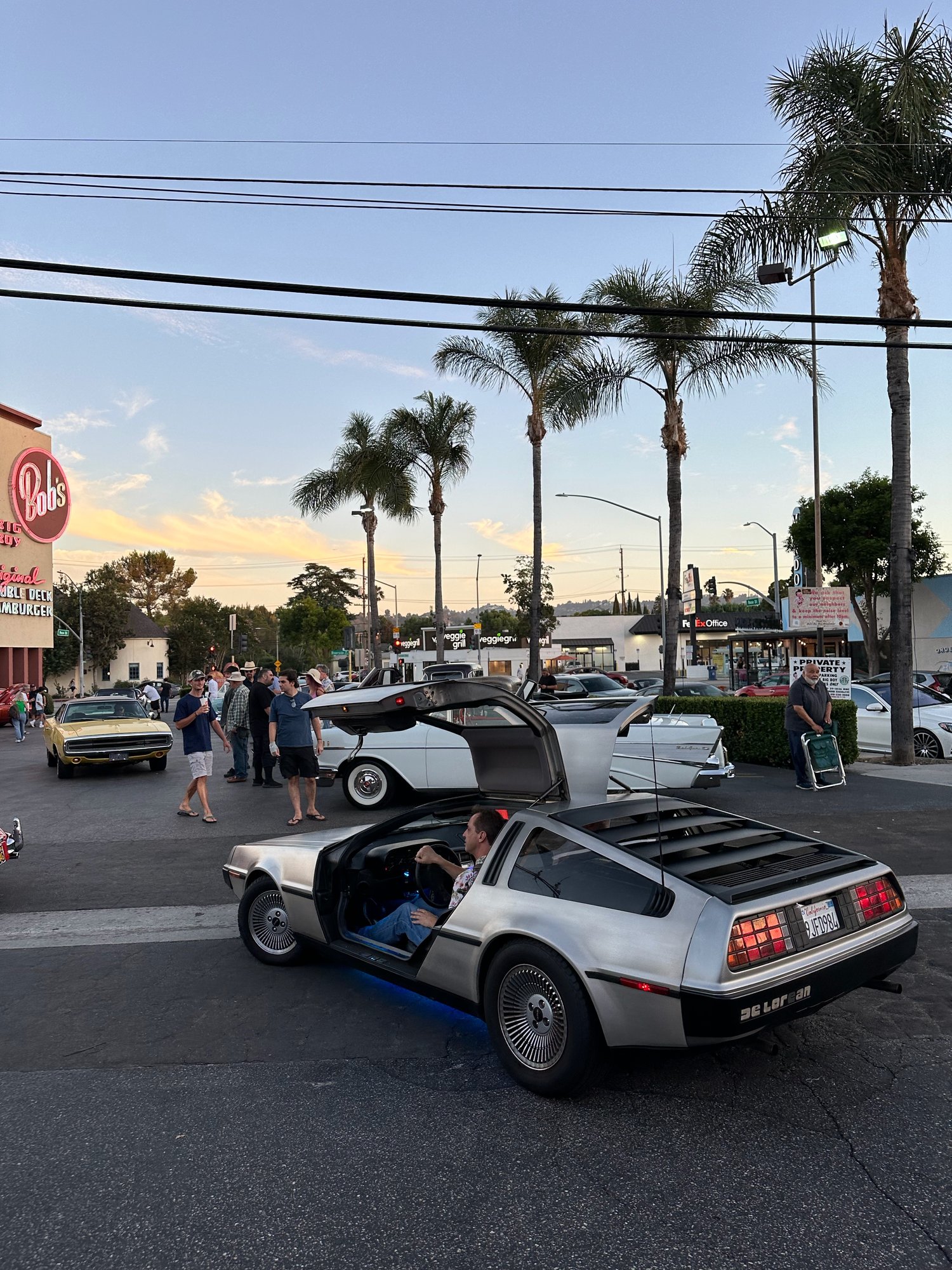 Silver DeLorean with gullwing door open at sunset beside the Bob's Big Boy sign in Burbank, crowd around
